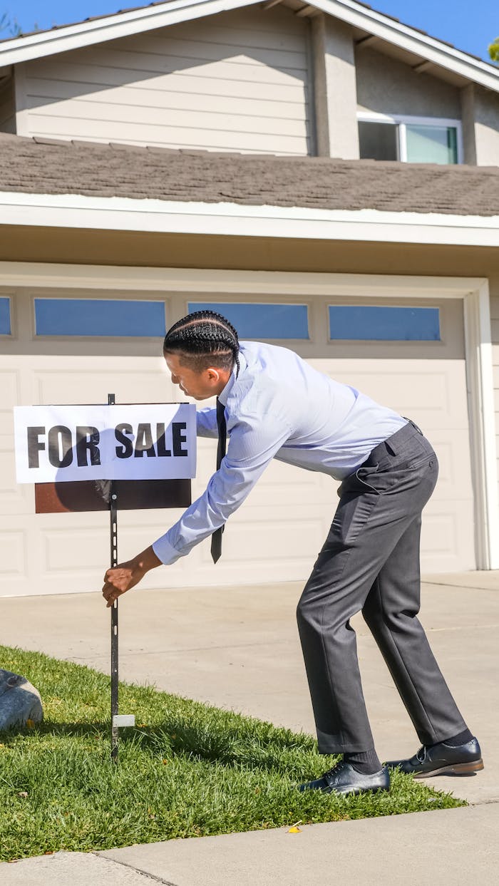 Black male realtor placing 'For Sale' sign in front of a suburban house.
